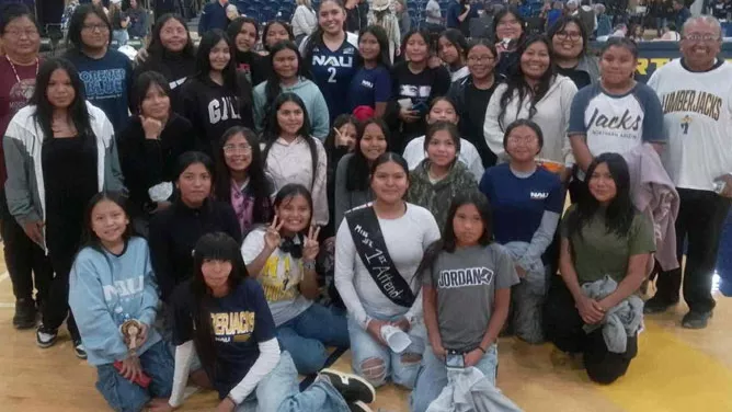 Large group of students and a few adults pose together on a basketball court inside a gymnasium. They are smiling and wearing casual clothing, including several shirts with NAU and school logos.