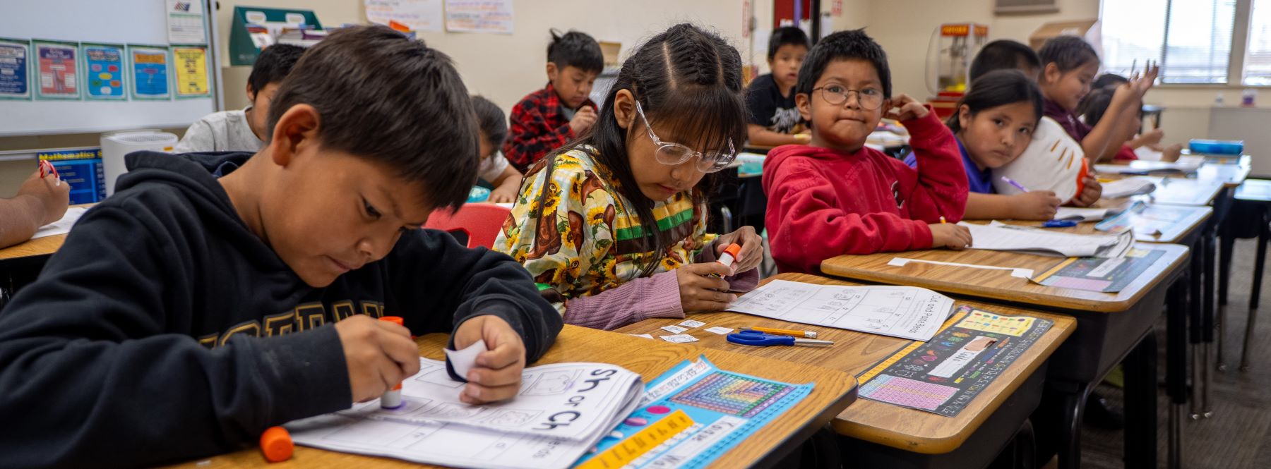 Students sitting at desks in a classroom, engaged in various school tasks with smiles and concentration on their faces.
