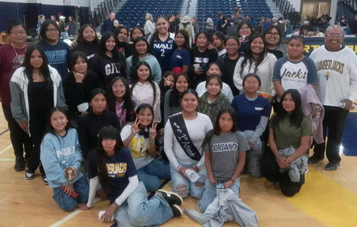 Large group of students and a few adults pose together on a basketball court inside a gymnasium. They are smiling and wearing casual clothing, including several shirts with NAU and school logos.