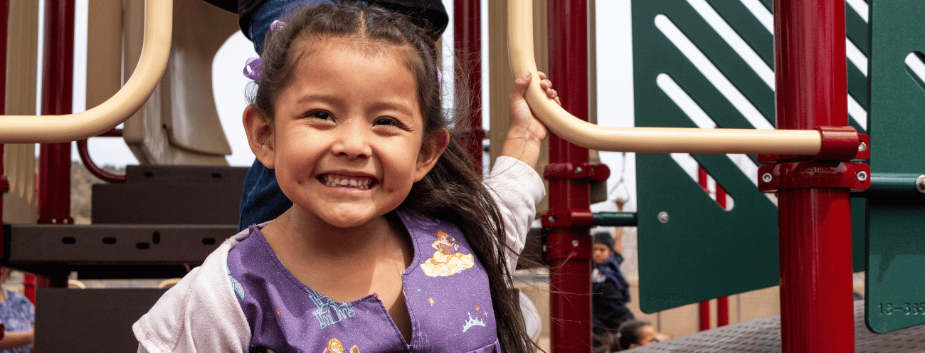 A smiling young girl playing on playground equipment, holding onto a handrail with one hand.
